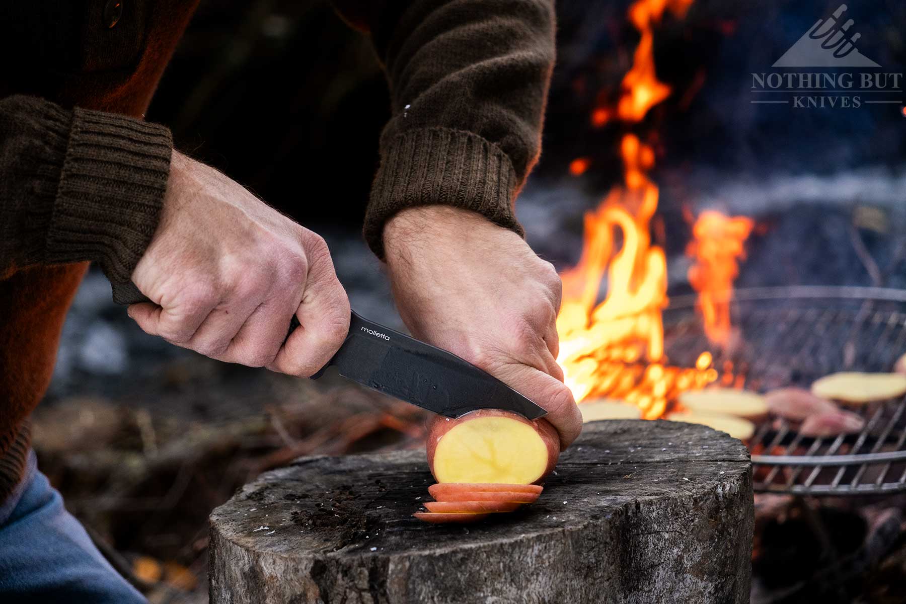 The LionSteel M5 being used to slice a potato for dinner in front of a campfire in the wilderness. 