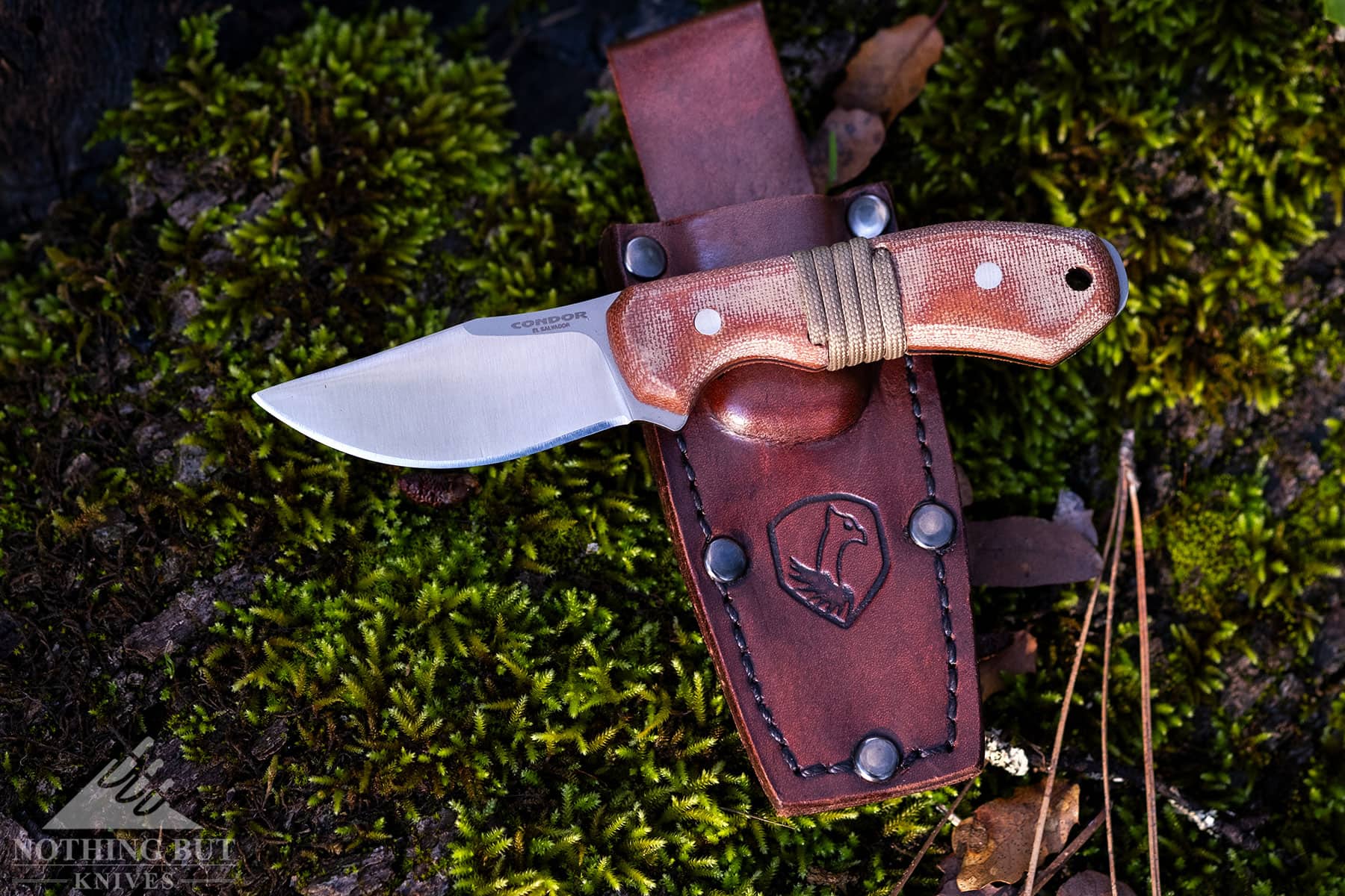 An overhead view of the Condor Mountaineer Trail Wingman knife laid across its brown, leather sheath on top of a mossy rock background. 