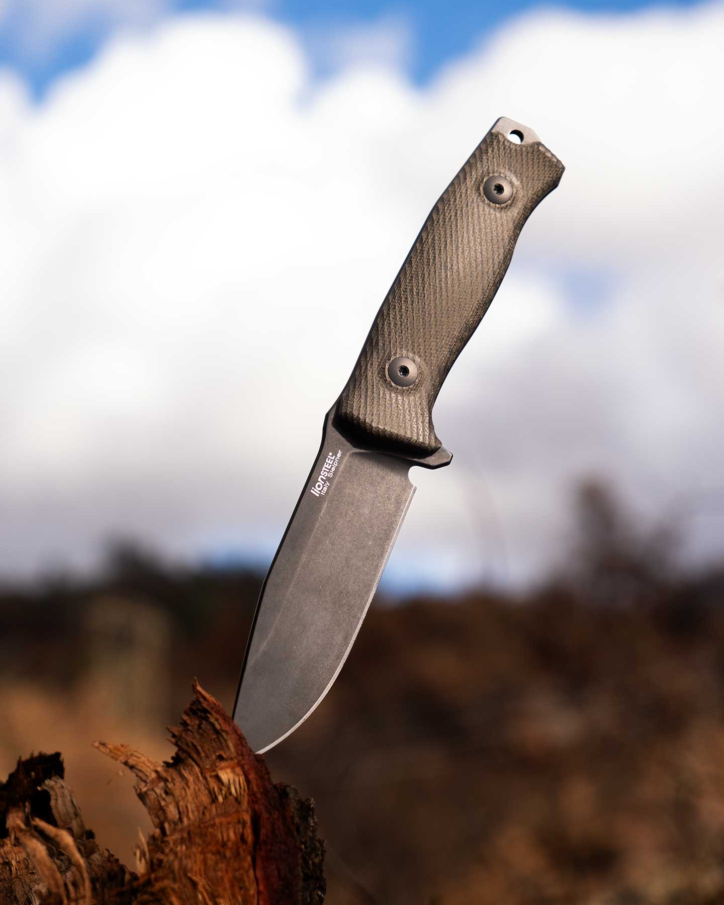 The LionSteel M5 survival knife with its blade tip stuck in a tree stump in front of a mountain ridge and a cloud filled blue sky.