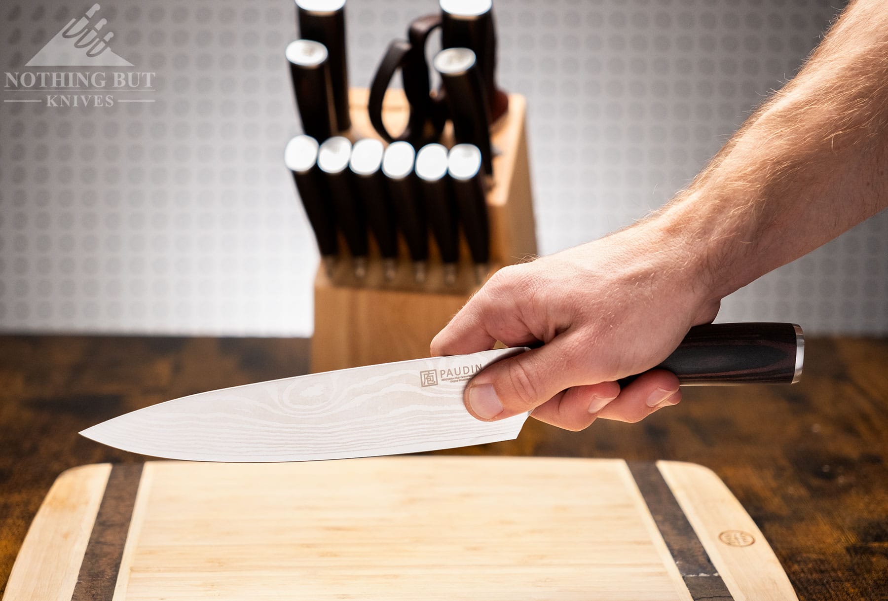 A close-up of a person holding a Paudin chef knife above a cutting board in front of the remainder of the knife set. 