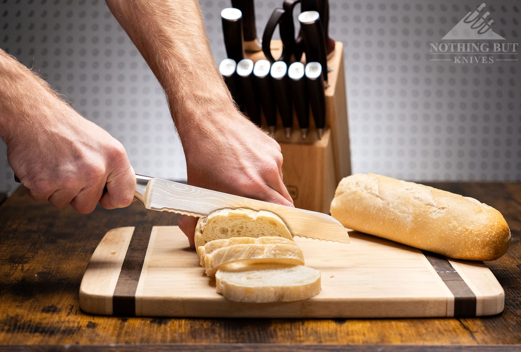 A Paudin 8-inch bread knife being used to slice a loaf of French bread with the rest of the Universal Collection knife set in the background. 