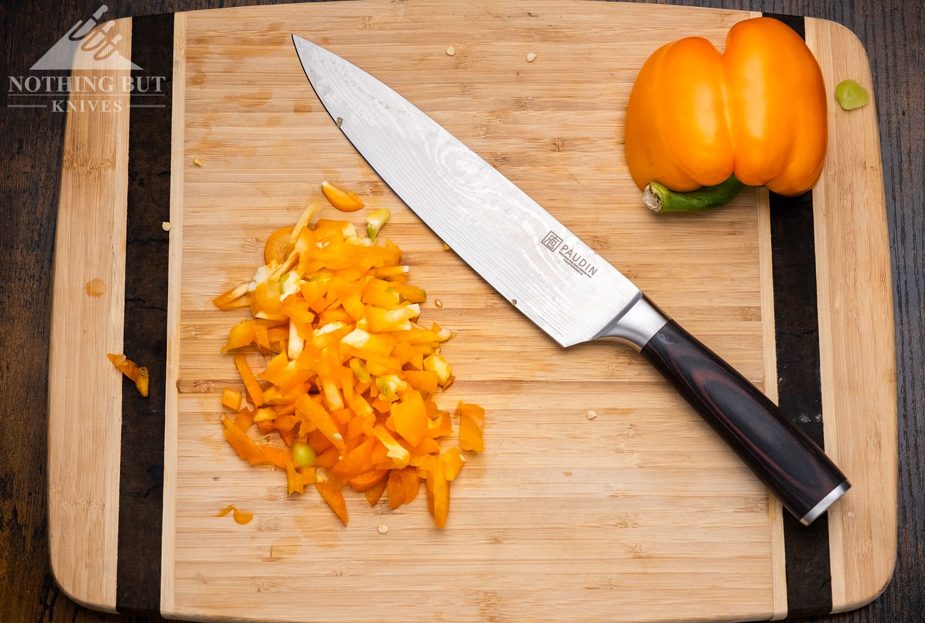 An overhead view of the Paudin Universal Collection chef knife laying on a bamboo wood cutting board next to a pile of diced yellow peppers it was used to cut.