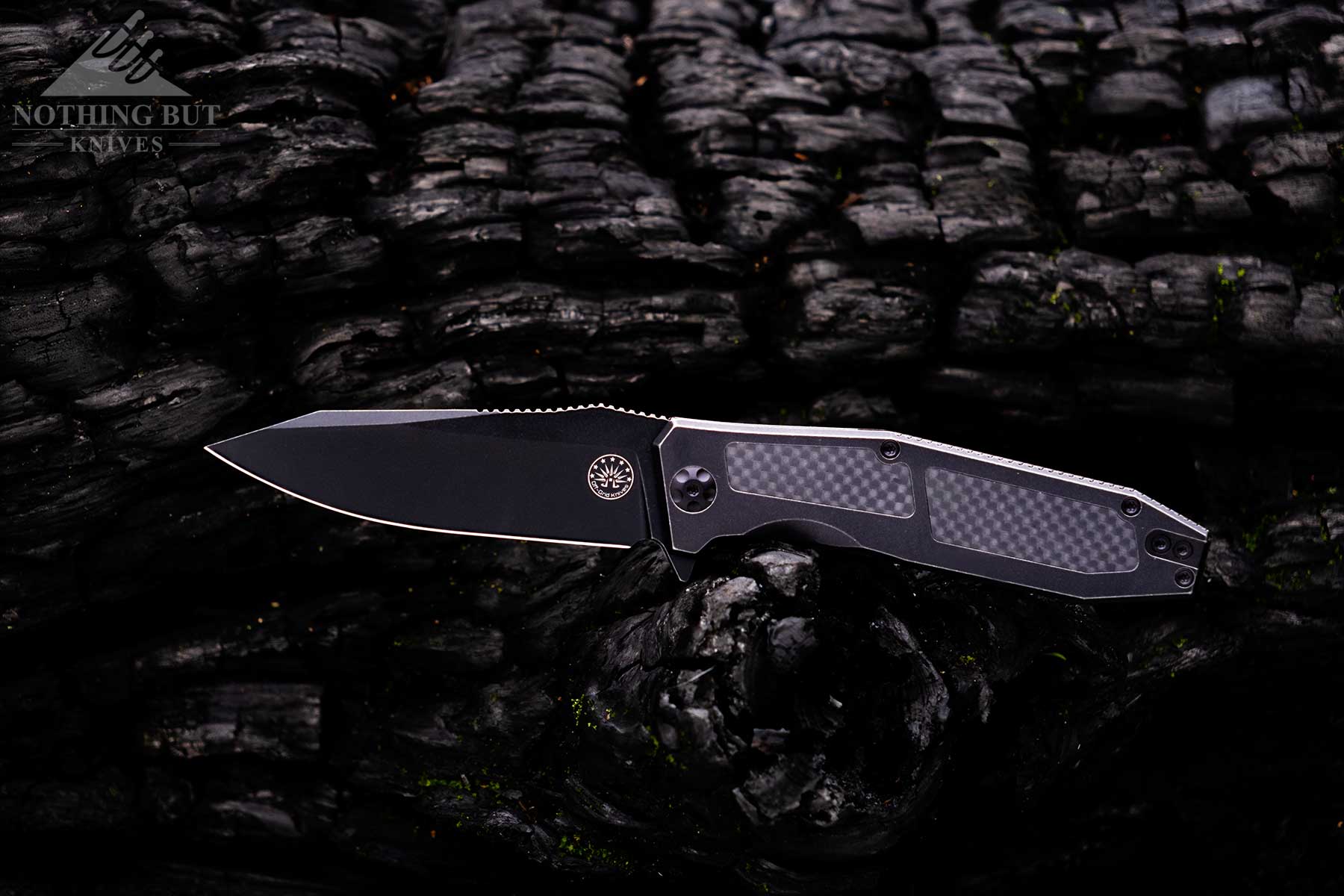 The Off-Grid Scorpion V3 folding knife with its blade in the open position balanced on a charred log. 