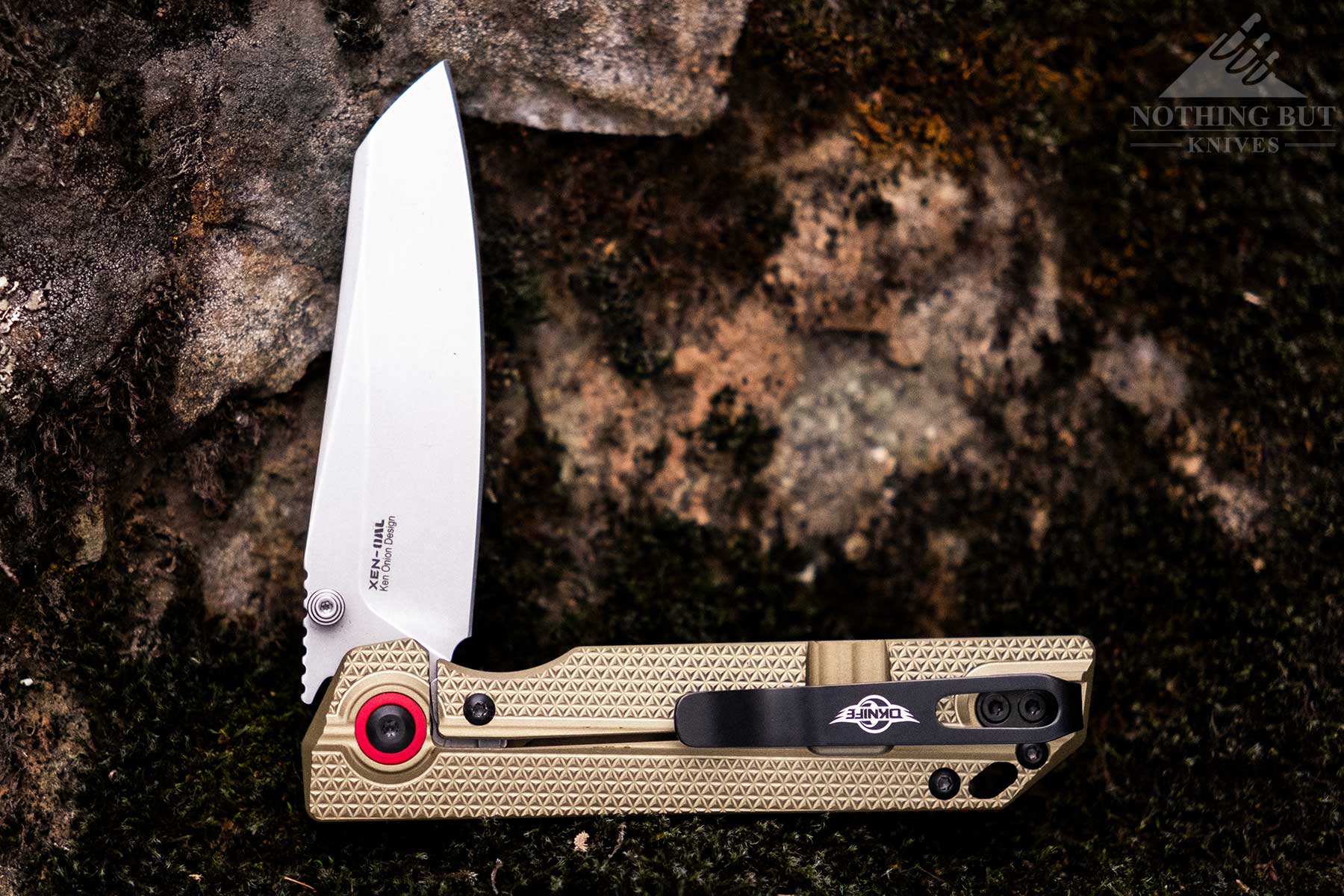 The Oknife XEN-OAL pocket knife on a moss-covered rock with its blade in the half open position. 