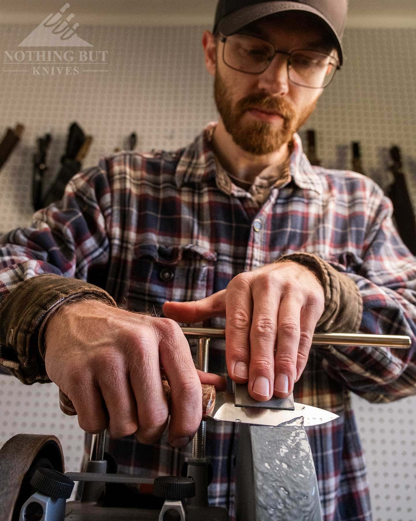 A low angle shot of a man man sharpening a knife on the Tormek T-4 grinder in front of a wall containing a variety of fixed blade knives.