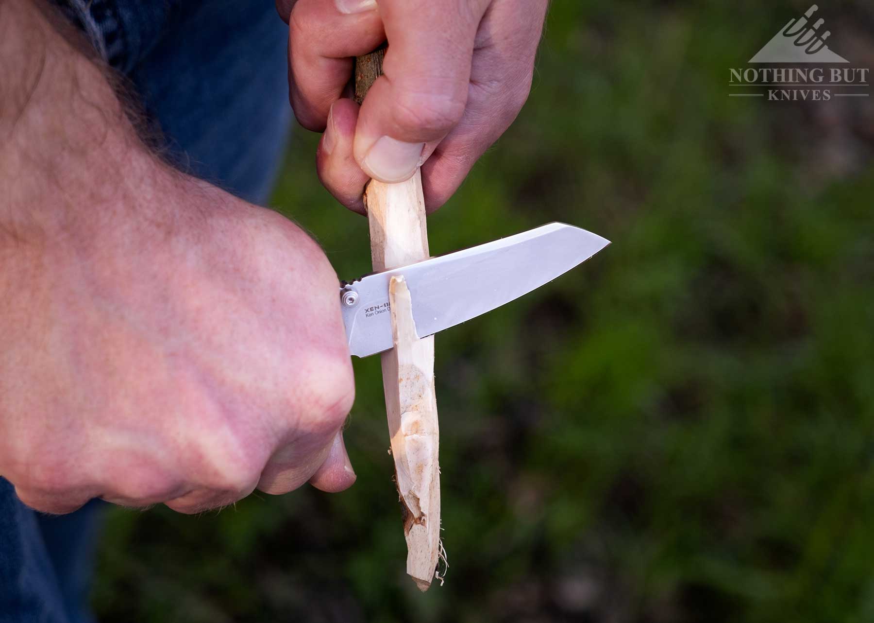 A close-up of the Oknife XEN-OAL folding knife being used to carve a stick in front of a green background. 