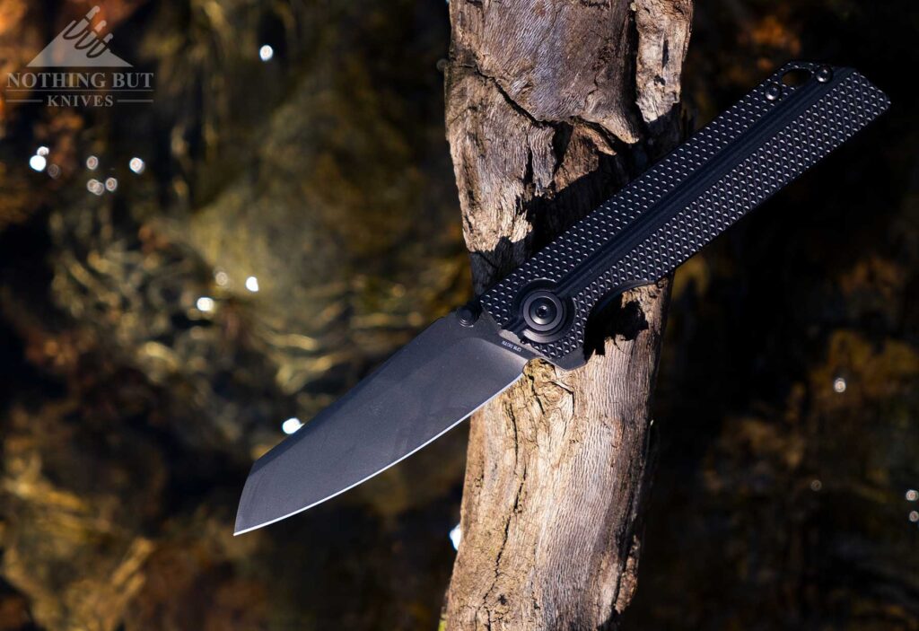 The matte black version of the XAN-AOL pocket knife with the blade in the open position on a log above a creek. 