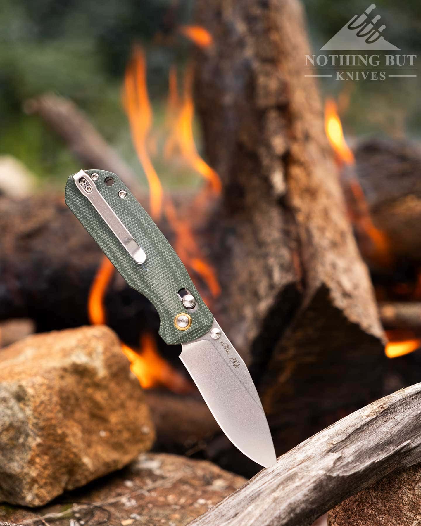 The CJRB Cutlery folding knife shown here with its blade in the open position sticking out of a piece of driftwood next to a campfire. 